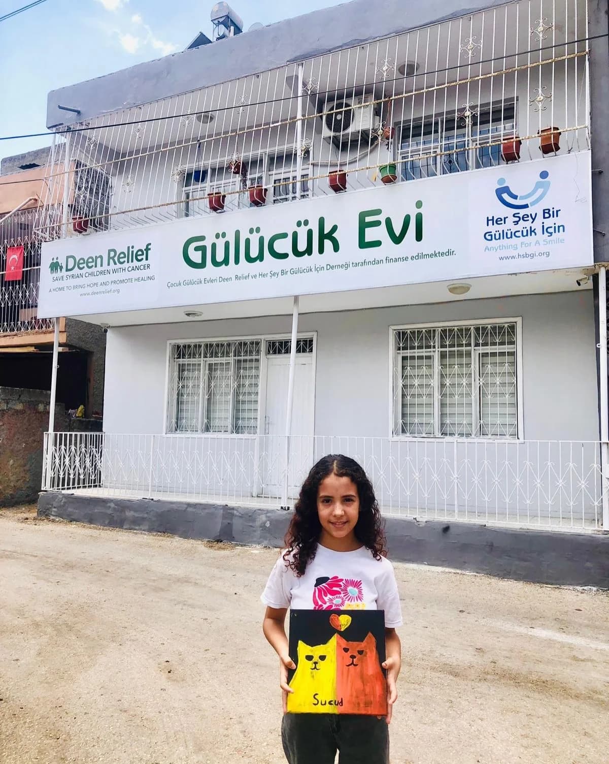 A young girl holding her painting in front of the Gülücük Evi care centre signage in Adana