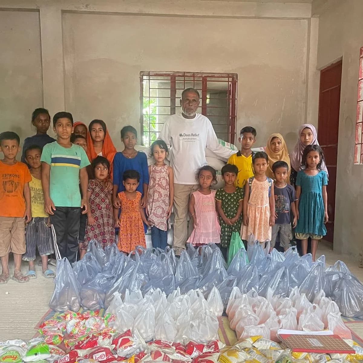 A Deen Relief field worker with around twenty Bangladeshi children, alongside dozens of packed Qurbani aid parcels and food supplies ready for distribution